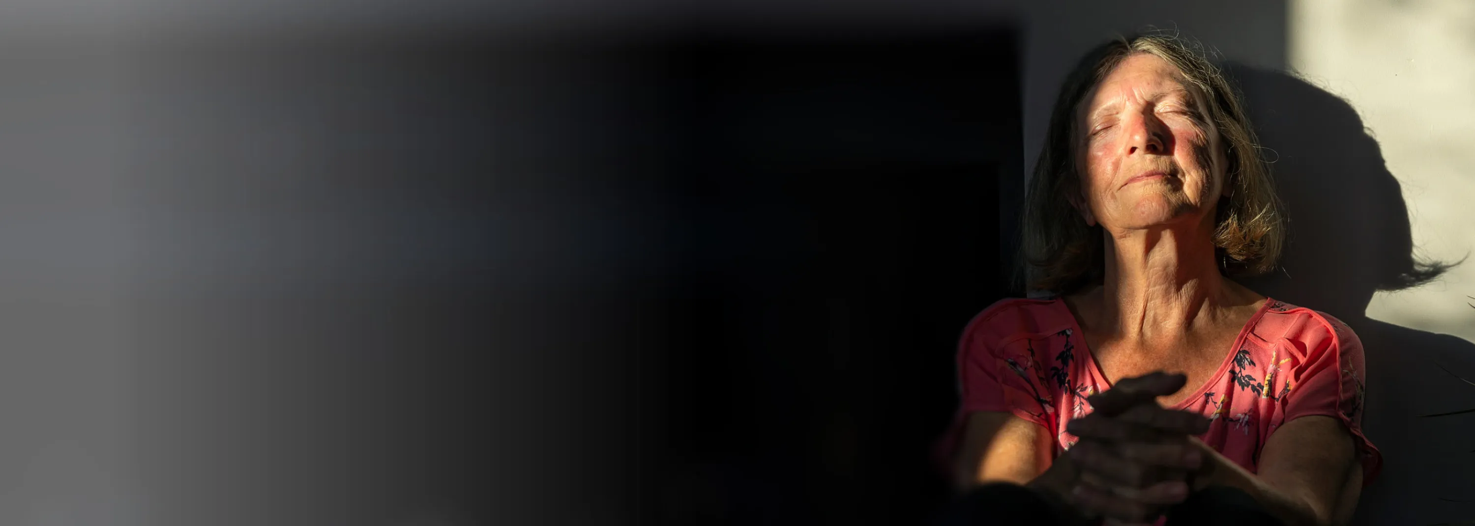 Une femme âgée est assise contre un mur, les yeux fermés, au soleil.